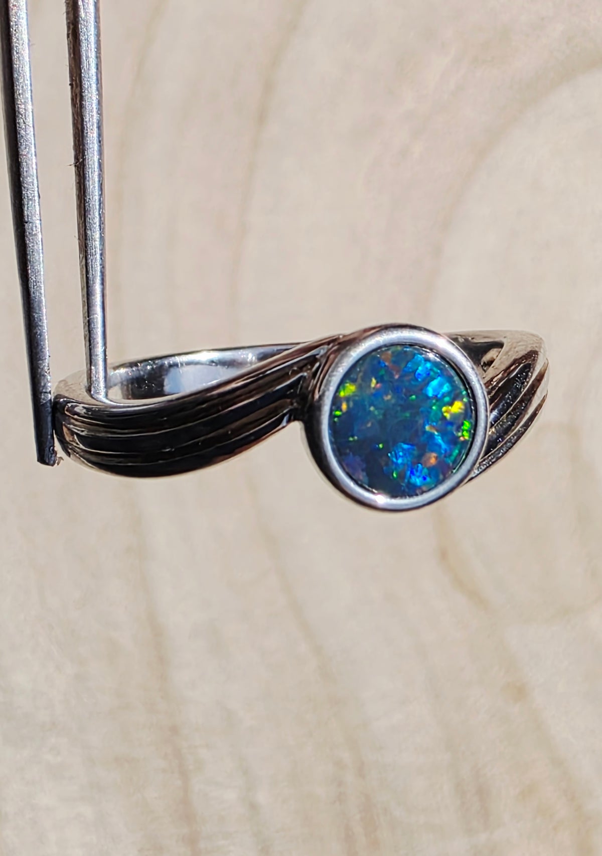 A silver ring with a round iridescent blue opal gemstone, held by metal tweezers against a light wood grain background.