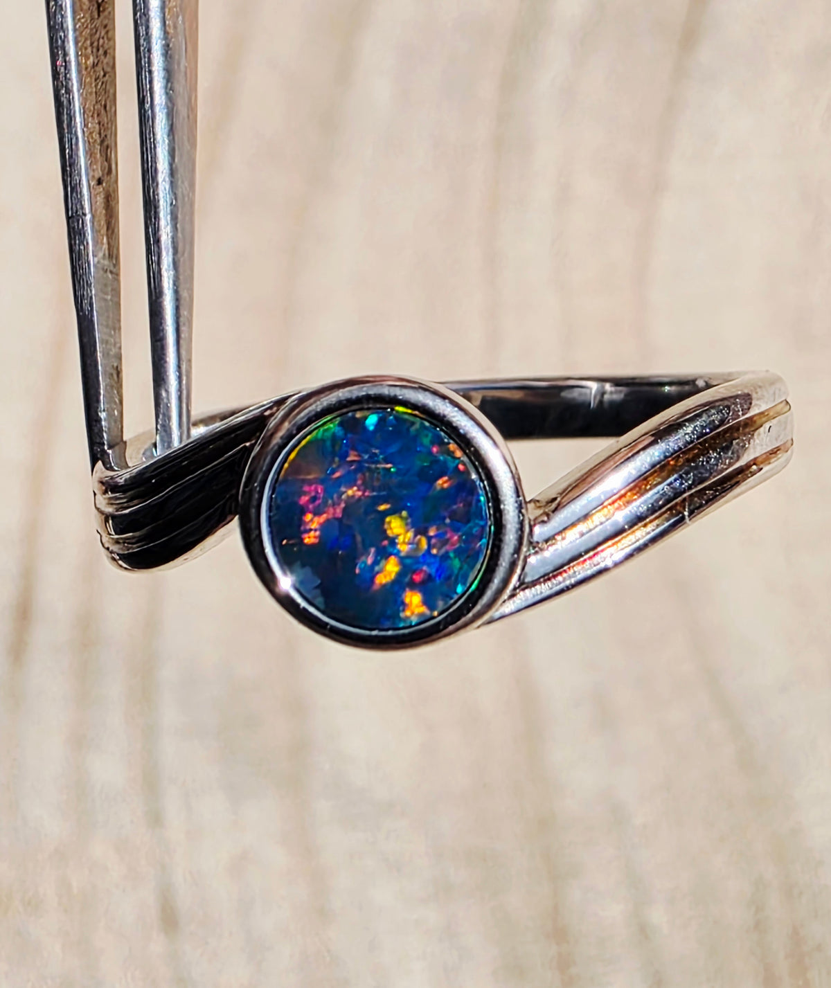 A silver ring with a round iridescent opal center, held by metal tweezers against a light wood grain background.
