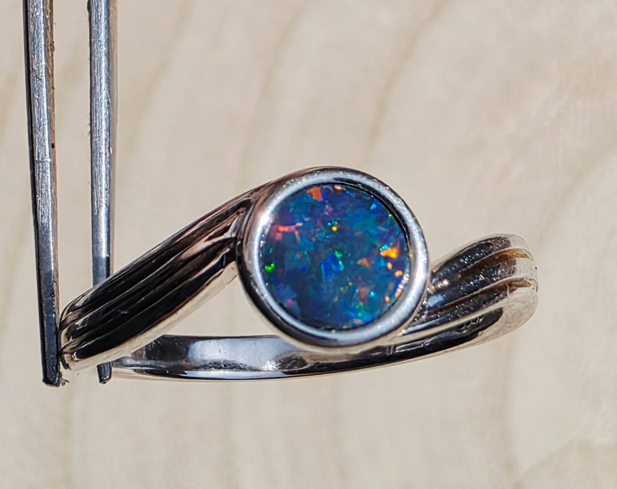 A silver ring with a round, vibrant blue opal gemstone, held in forceps against a light beige textured background.