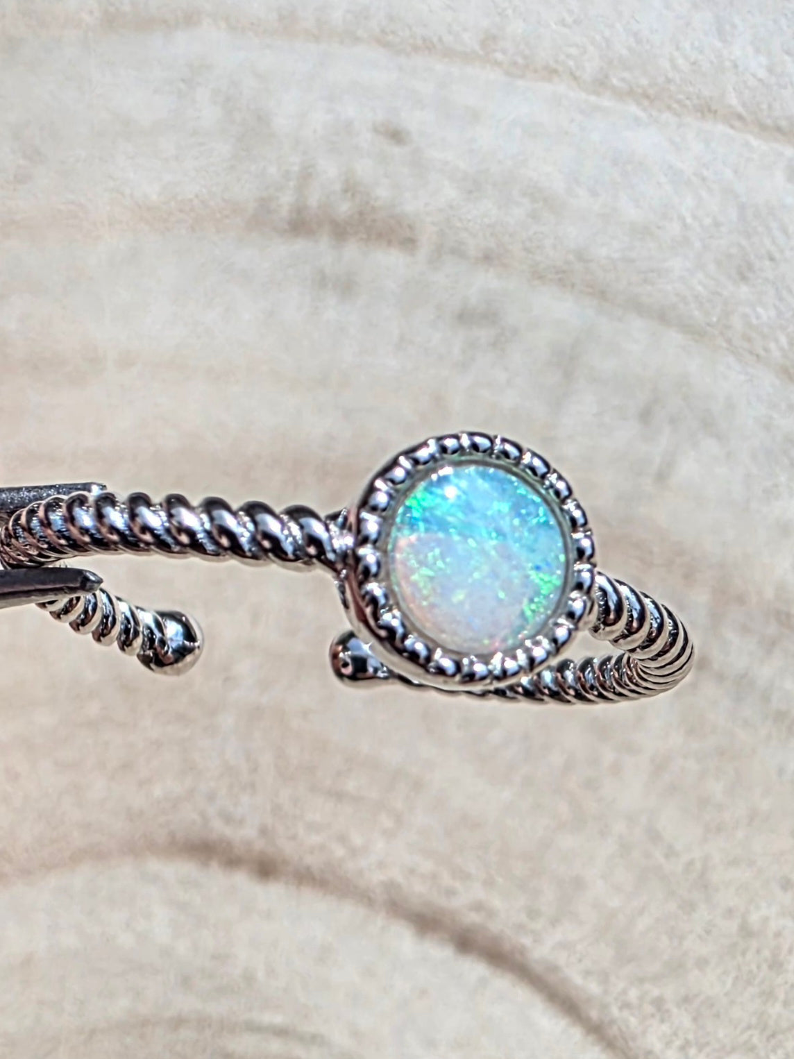 A silver twisted wire ring with a round iridescent white opal centre stone, photographed close-up against a soft beige textured background.