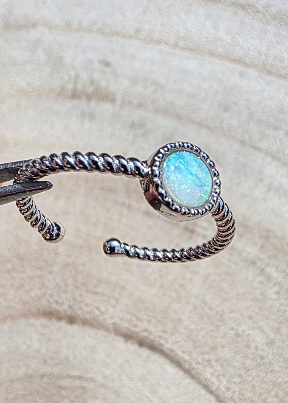 A silver twisted wire ring with a round iridescent white opal centre stone, photographed close-up against a soft beige textured background.