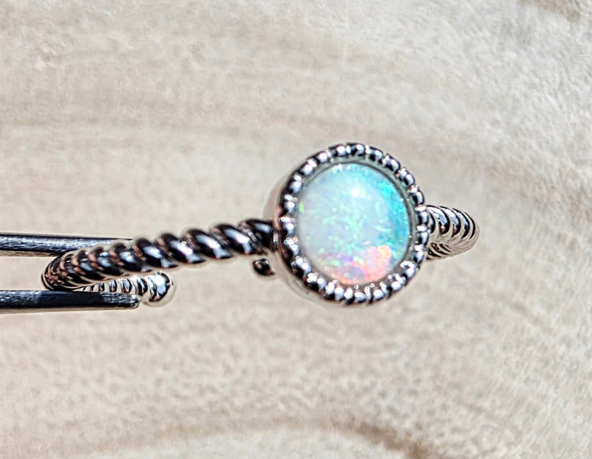 A close-up of a silver-toned ring with a round iridescent opal gemstone, held against a soft beige textured background.
