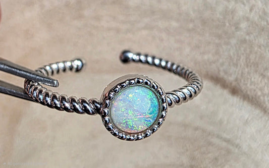A silver open ring  with a round iridescent opal centre stone, held by metal tweezers against a soft beige fabric background.