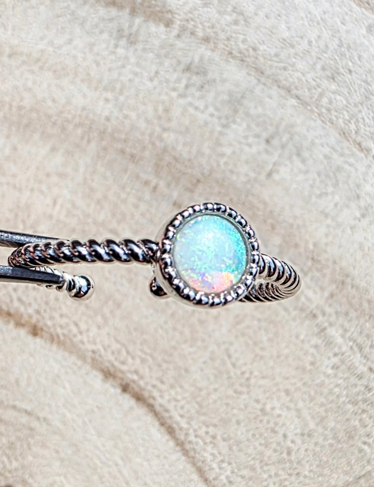 A silver twisted rope bangle ring featuring a round iridescent opal stone in a beaded silver bezel, displayed on a textured beige fabric background.