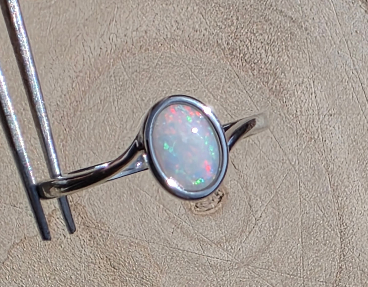 A sterling silver ring with an oval-cut white opal showing flashes of green and pink, held by metal tweezers against a textured beige background.