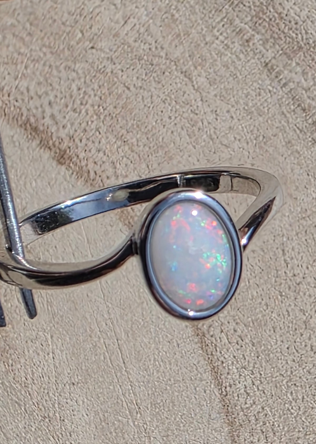 A silver ring featuring an oval white opal gemstone with vibrant multicolour flashes, displayed on a textured beige surface.