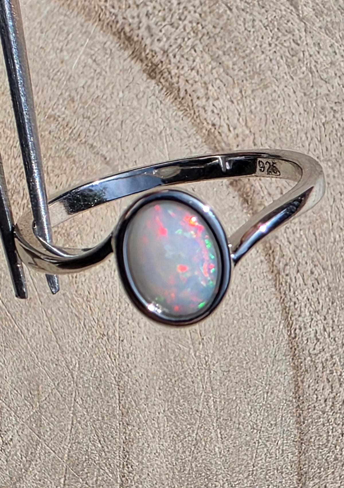 A sterling silver ring with an oval white opal centre stone, held by metal tweezers against a textured beige background. The inside of the band is stamped with '925'.