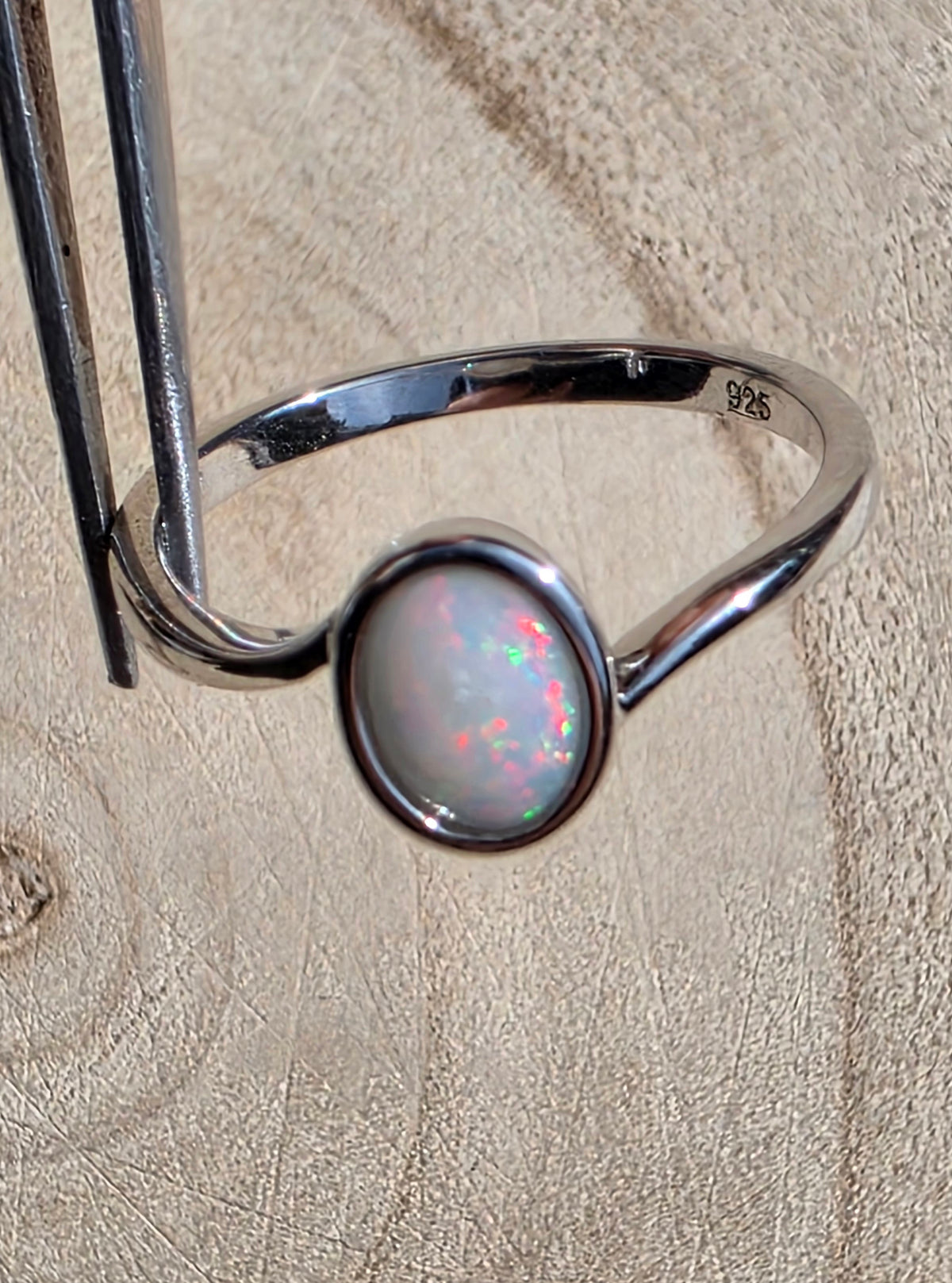 A sterling silver ring with an oval cabochon opal gemstone held by metal tweezers against a textured beige background. The opal displays a play-of-colour with green, pink, red and blue flashes.
