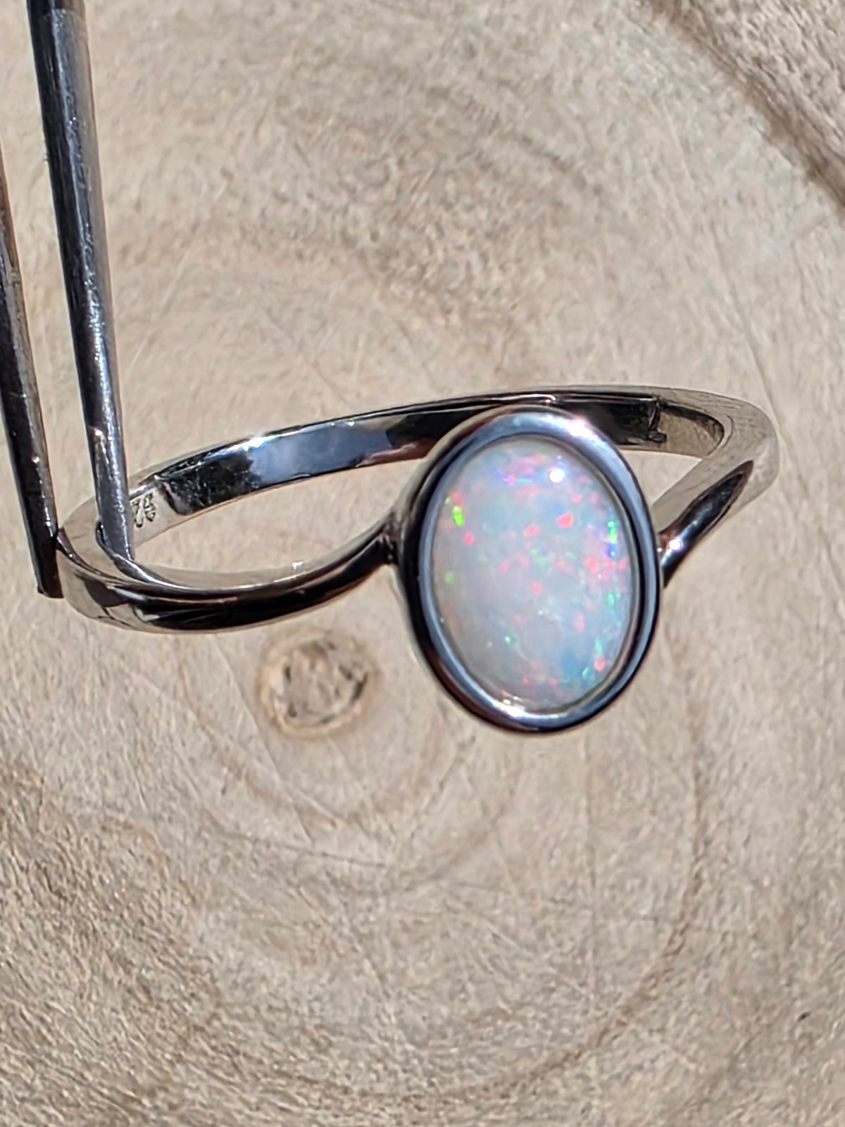 A silver ring with an oval cabochon opal gemstone, held by metal tweezers against a textured light wood background.