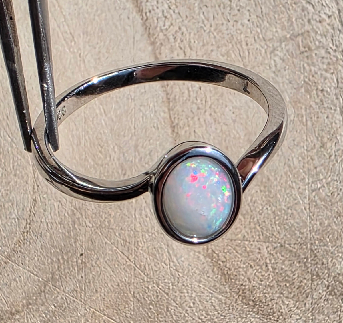 A sterling silver ring with a round bezel-set white opal showing iridescent pink and blue flashes, held by forceps against a textured beige background.