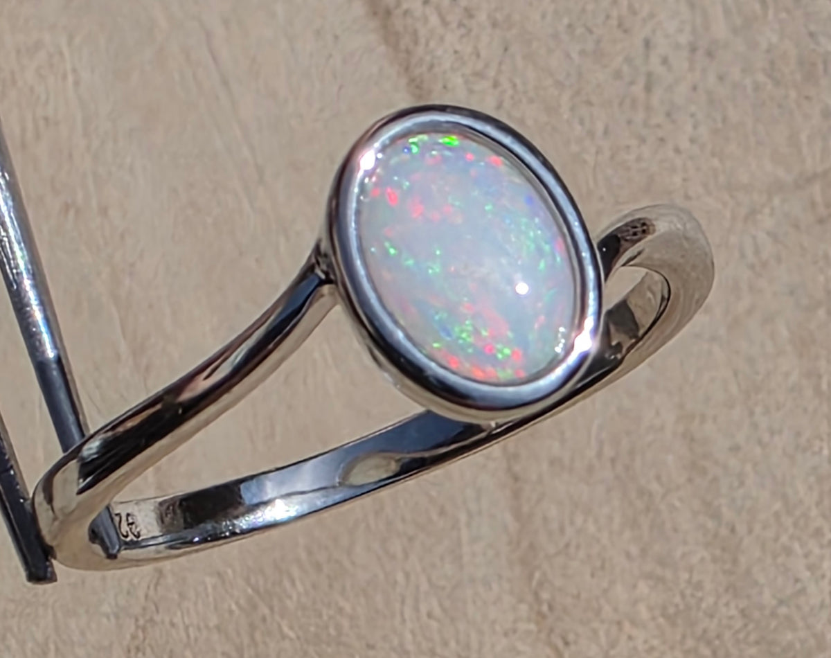 A sterling silver ring featuring an oval cabochon opal with vibrant green, pink, and blue flashes, displayed on a stand against a textured beige background.
