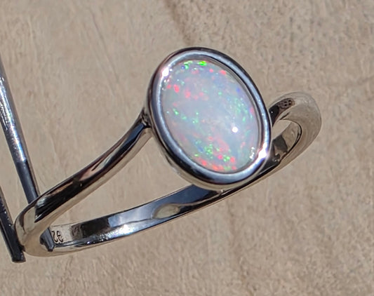 A sterling silver ring featuring an oval cabochon opal with vibrant green, pink, and blue flashes, displayed on a stand against a textured beige background.
