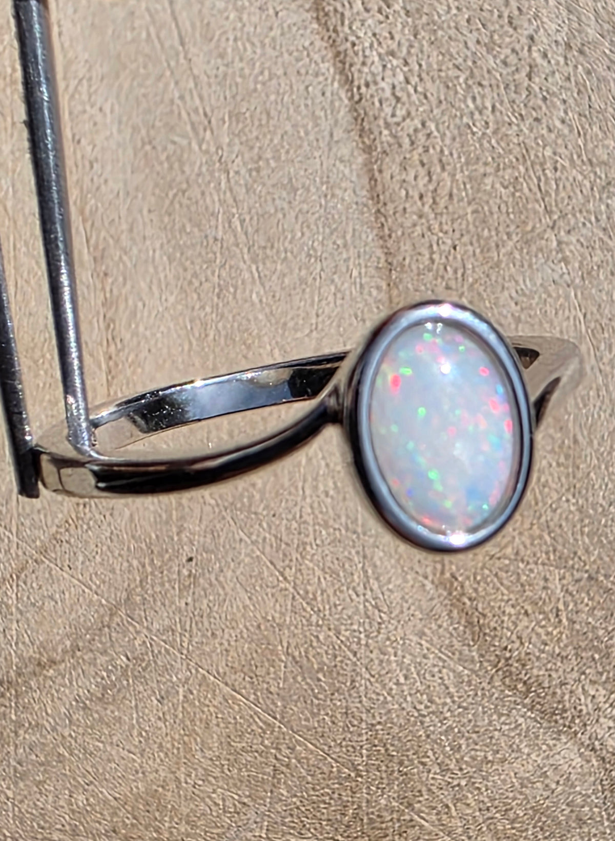 A silver ring featuring an oval-shaped white opal gemstone with iridescent pink, green, and blue flashes, displayed on a textured beige surface.