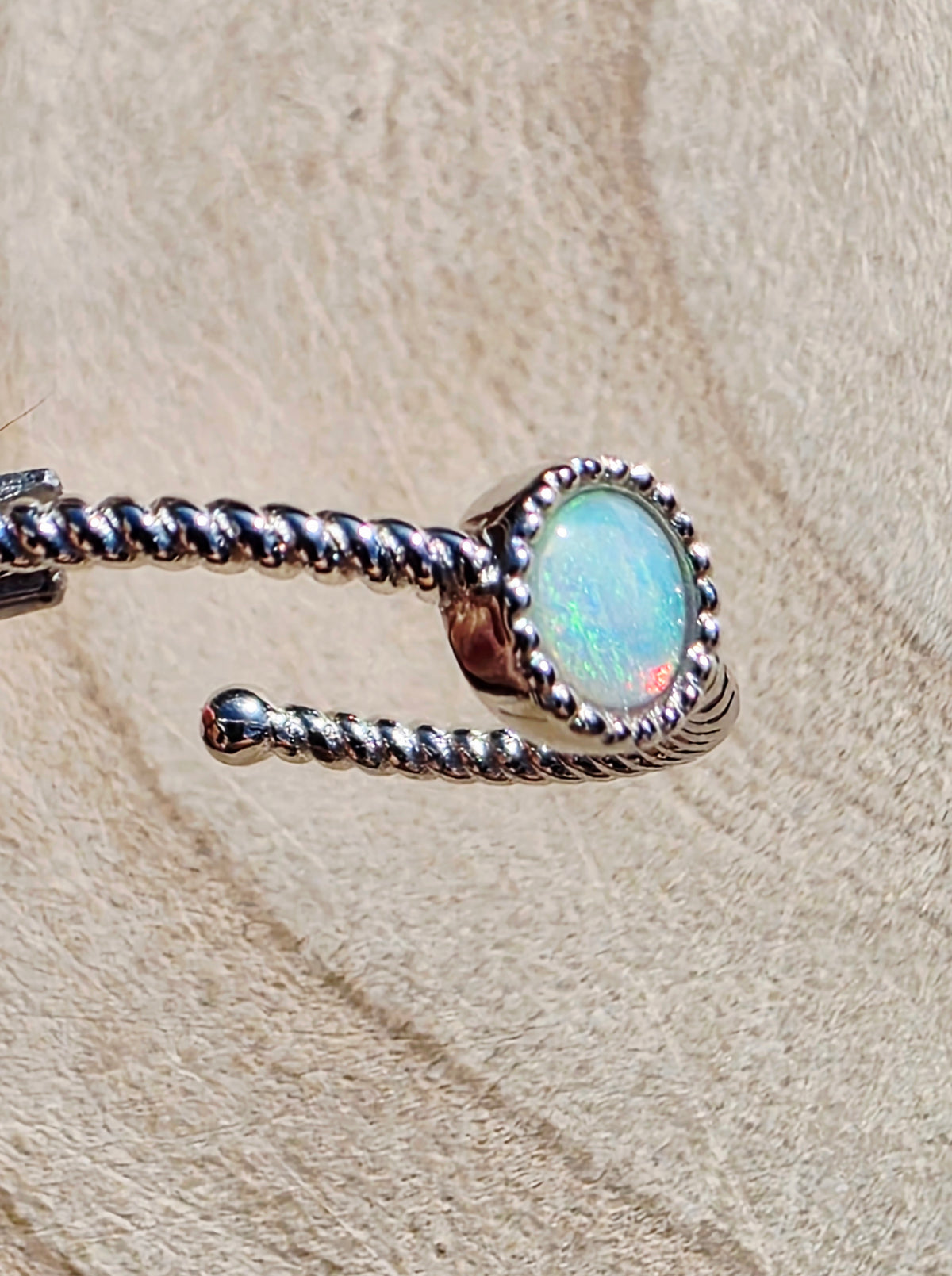 A silver-toned twisted rope ring with an oval iridescent opal stone, displayed on a textured beige surface.