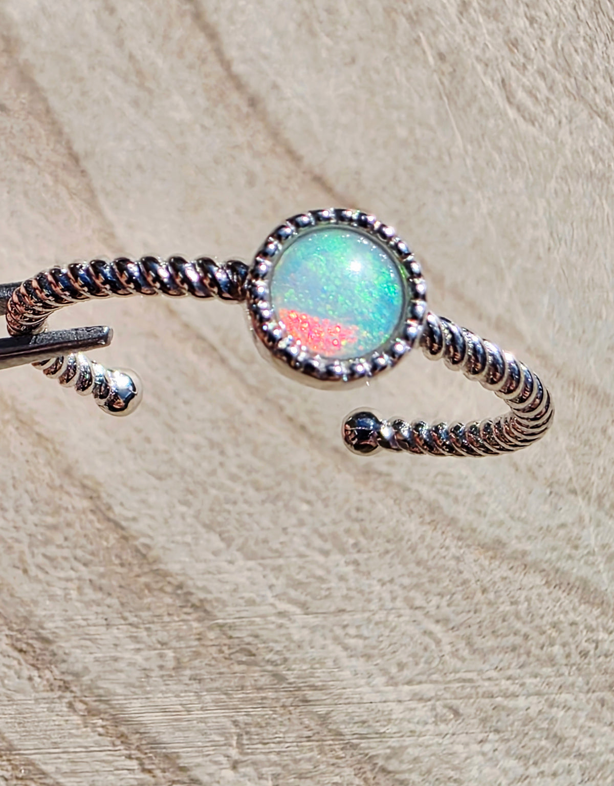 A silver twisted open ring featuring a round bezel-set iridescent opal stone with green, blue, and pink flashes, displayed on a textured beige fabric background.