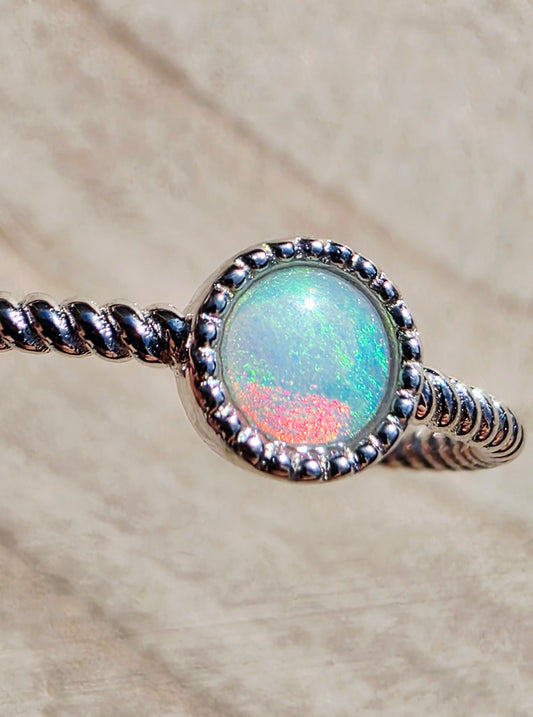 A silver-toned twisted wire ring featuring a round bezel-set opal stone with iridescent green, blue, and pink flashes, shown close-up on a textured beige background.