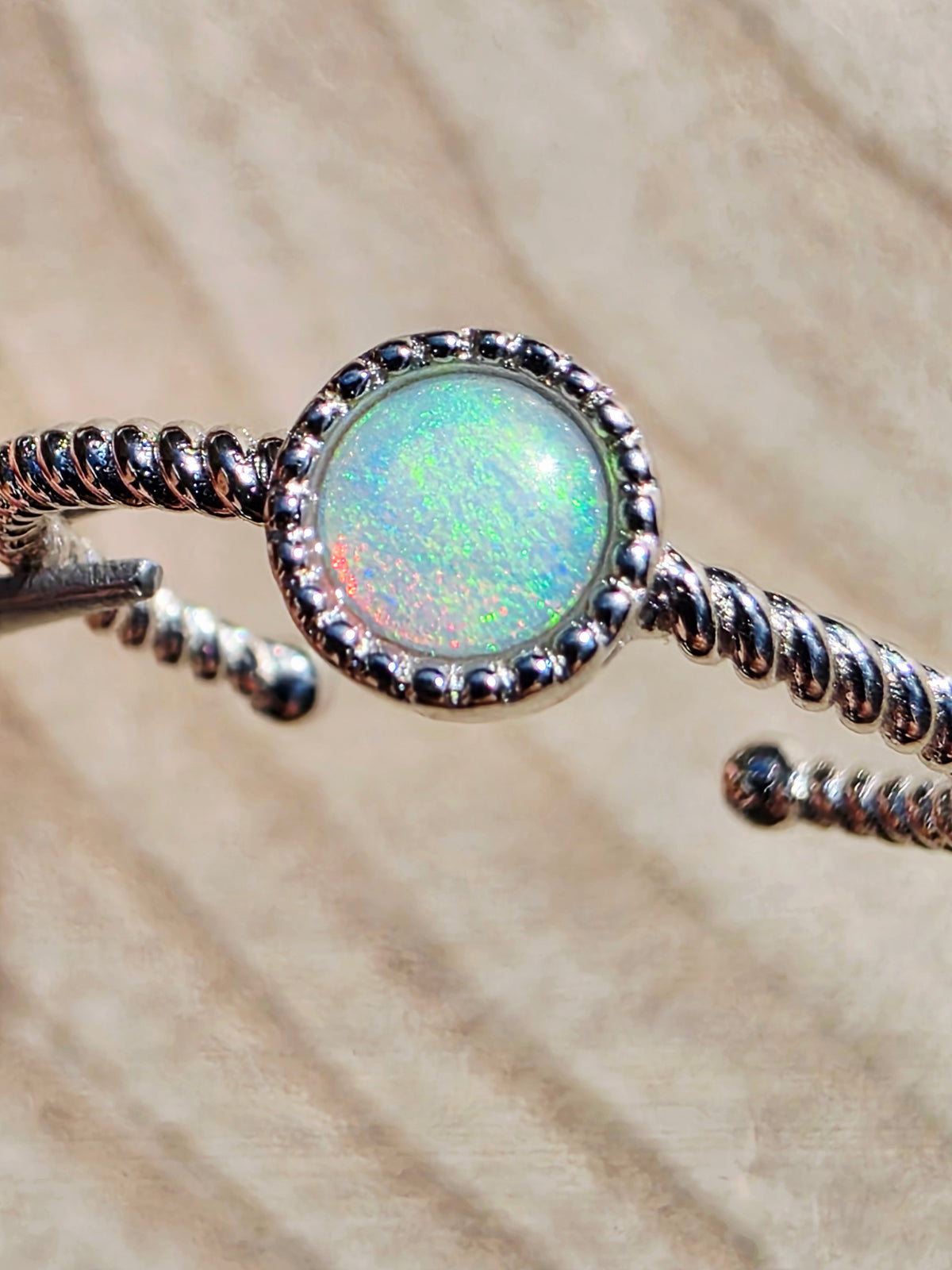 A close-up of a silver twisted wire cuff ring featuring a round iridescent opal stone in the centre, set against a textured beige background.