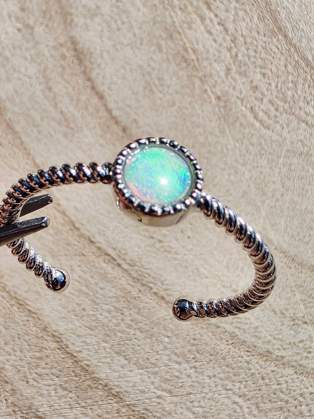A silver open cuff ring featuring a round iridescent opal stone in the centre, resting on a textured beige background.