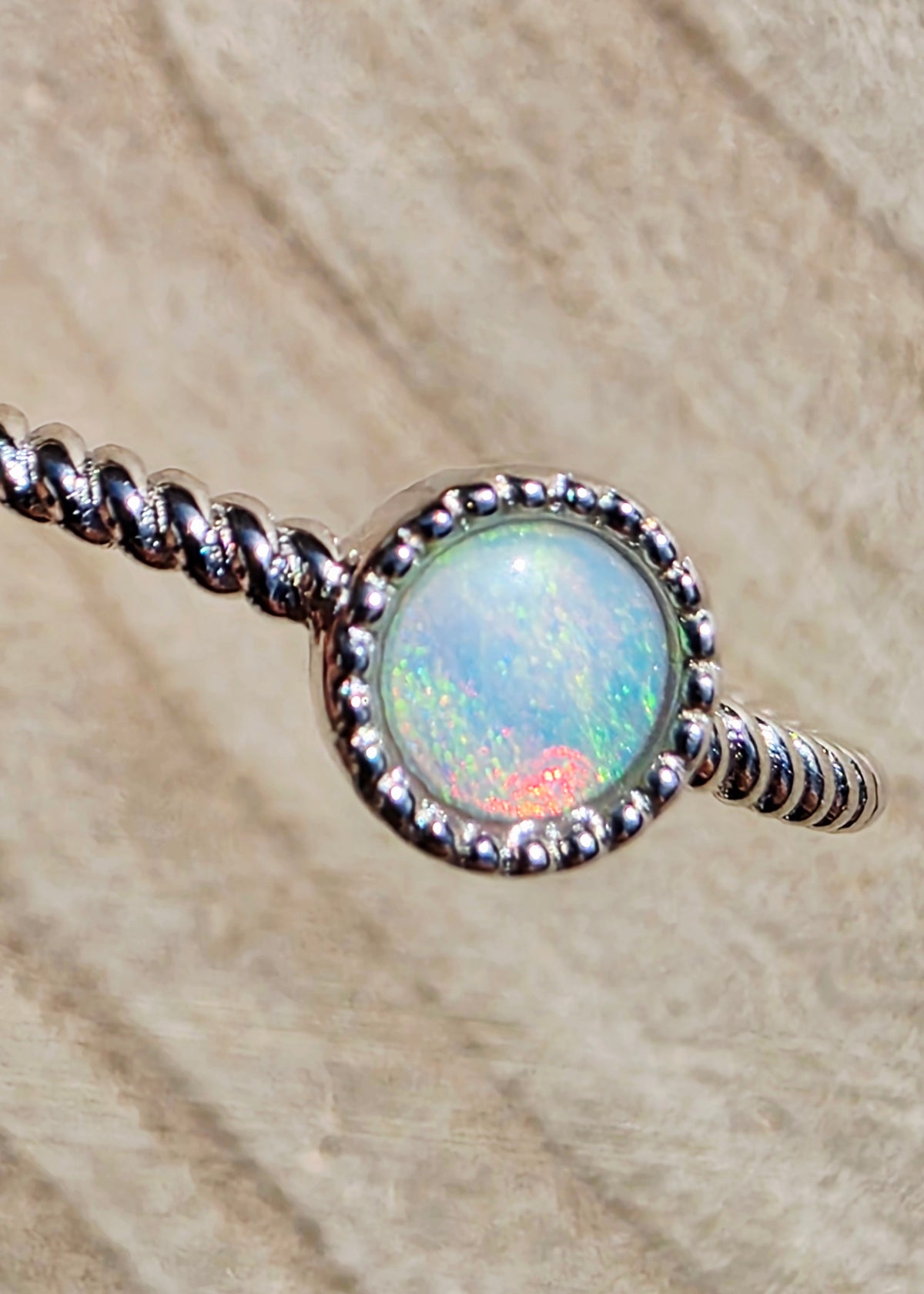 A silver twisted wire ring featuring a round bezel-set iridescent opal stone with blue, green, and orange flashes, shown against a textured beige background.