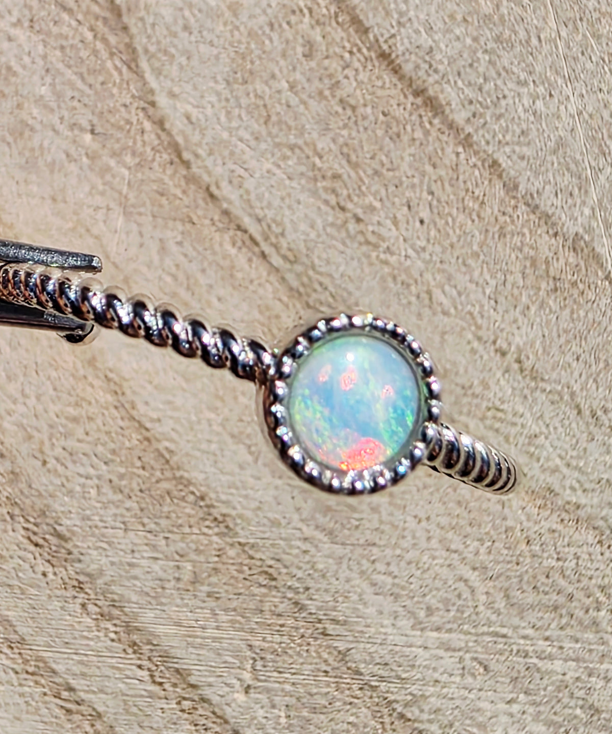 A silver-toned twisted band ring with a round iridescent opal centre stone, photographed close-up on a textured beige surface.