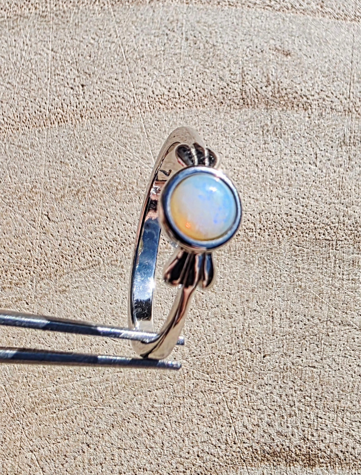 A sterling silver ring with a round white opal gemstone, held in forceps against a textured beige background.