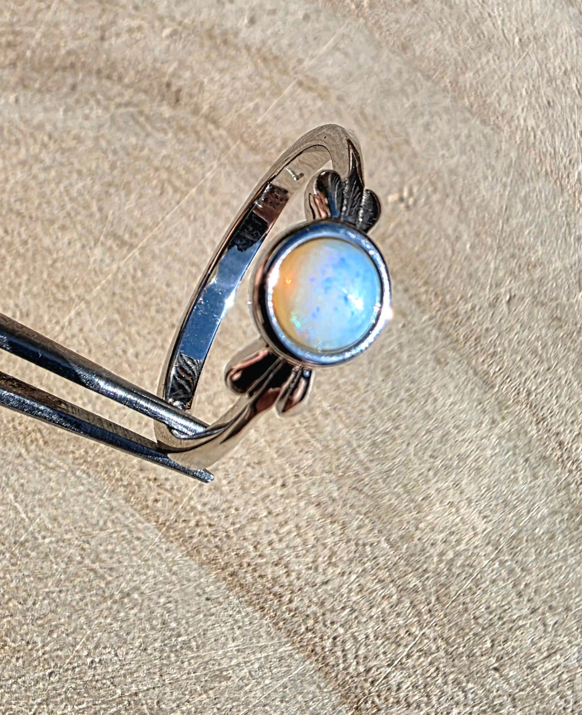 A sterling silver ring with a round iridescent opal cabochon, held in forceps against a textured beige background.