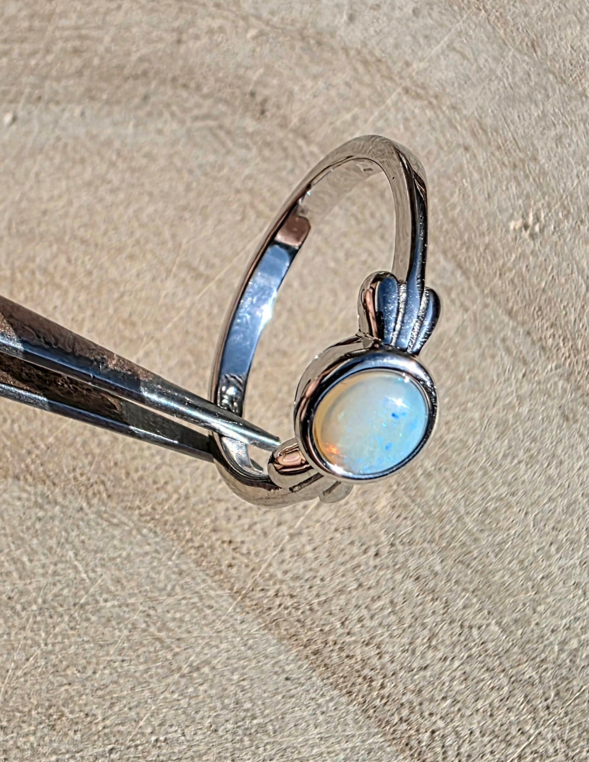 A sterling silver ring with an oval iridescent white cabochon stone, held in forceps against a textured beige background.