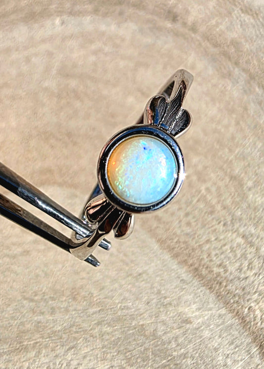 A sterling silver ring with a round iridescent opal gemstone, held in forceps against a textured beige background.