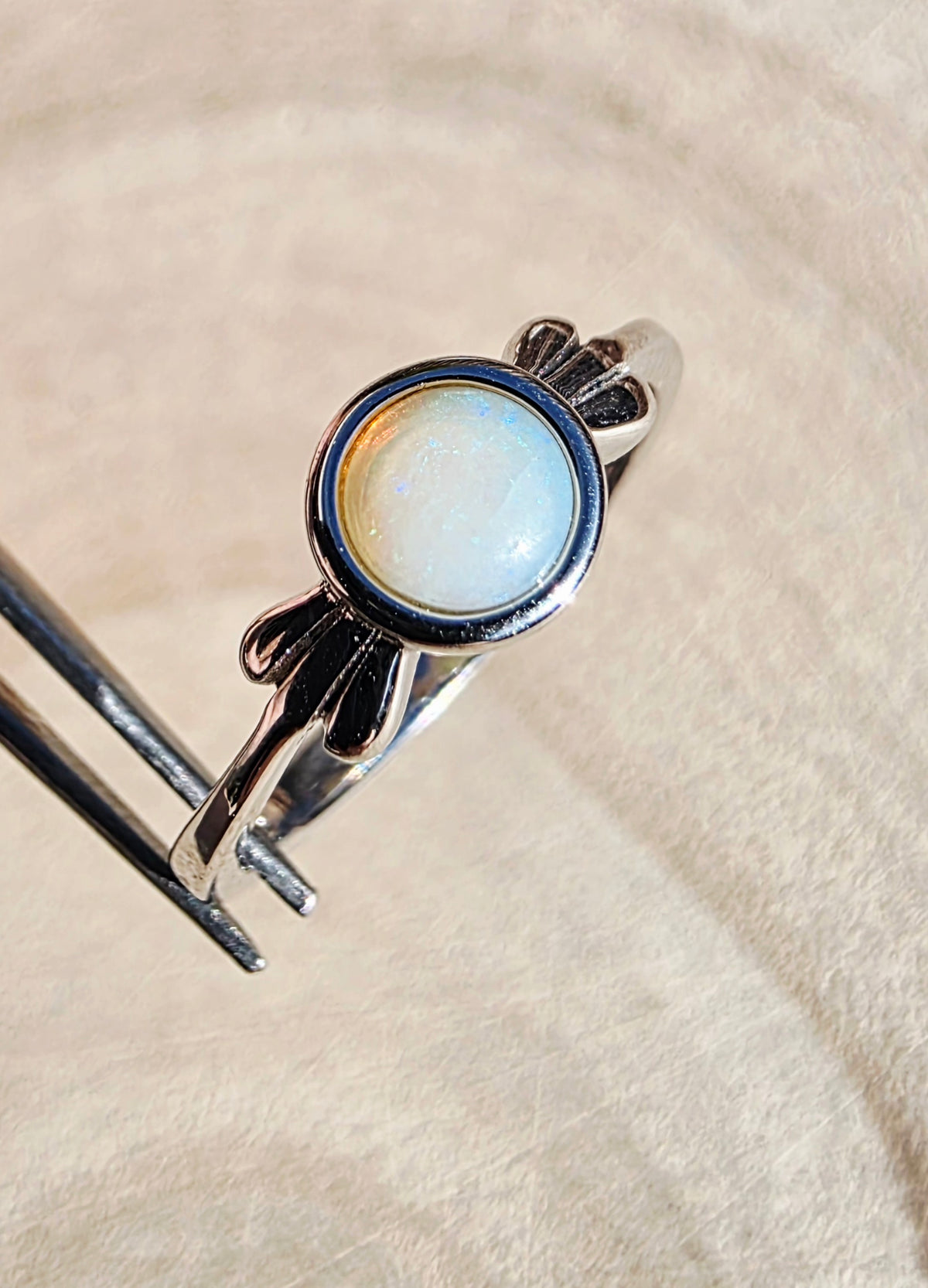 A silver ring with a round iridescent white cabochon gemstone, held by metal tweezers against a soft beige textured fabric background.