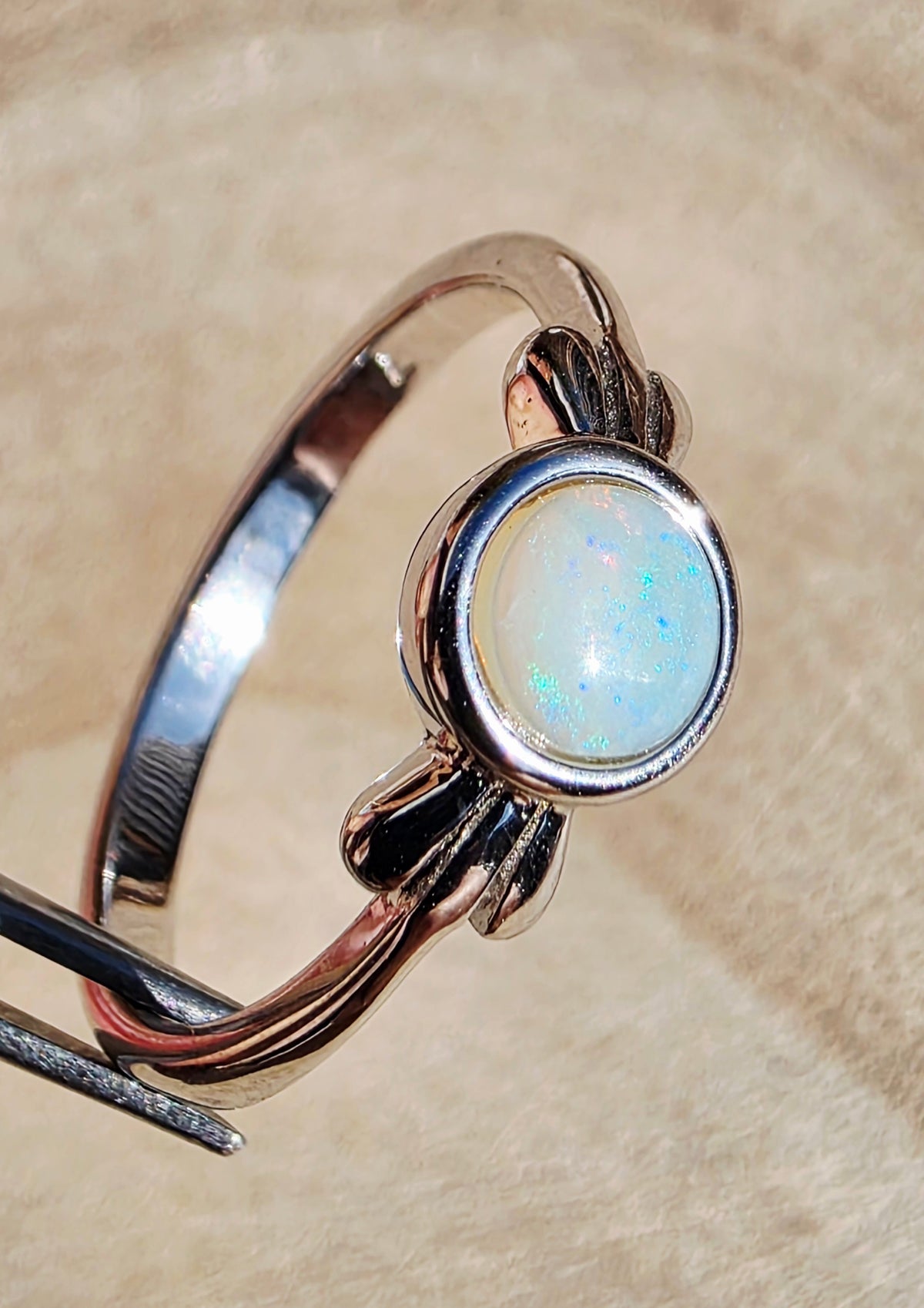 A silver-toned ring with a round iridescent white opal gemstone, held in place by a black stand against a soft beige textured background.