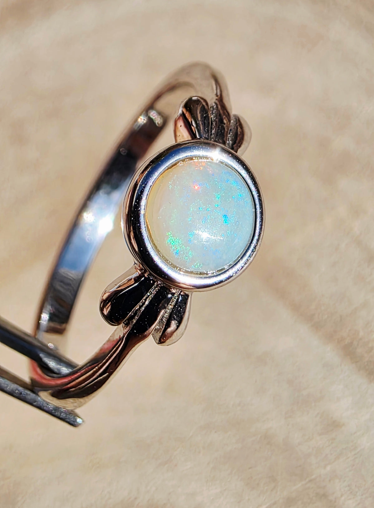 A sterling silver ring with a round white opal cabochon, held in forceps against a soft beige background.