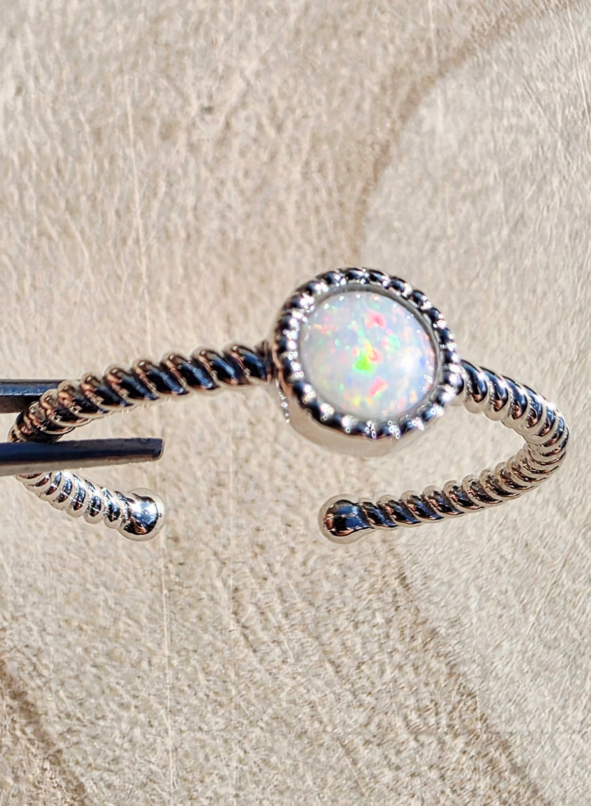 A silver-toned twisted open cuff ring featuring a round white opal center stone with iridescent flashes, photographed close up on a textured beige surface.
