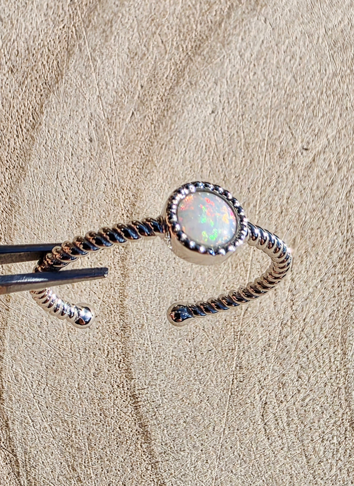 A silver twisted open ring with an oval iridescent opal centre stone, held by metal tweezers against a textured beige background.