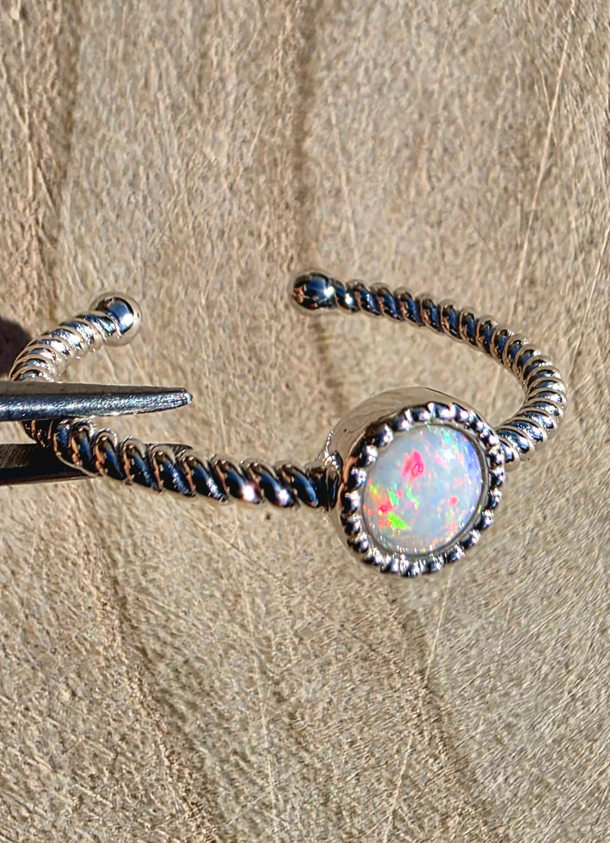 A silver-toned twisted wire open cuff ring featuring a round white opal stone with multicoloured flashes, displayed on a textured beige surface.