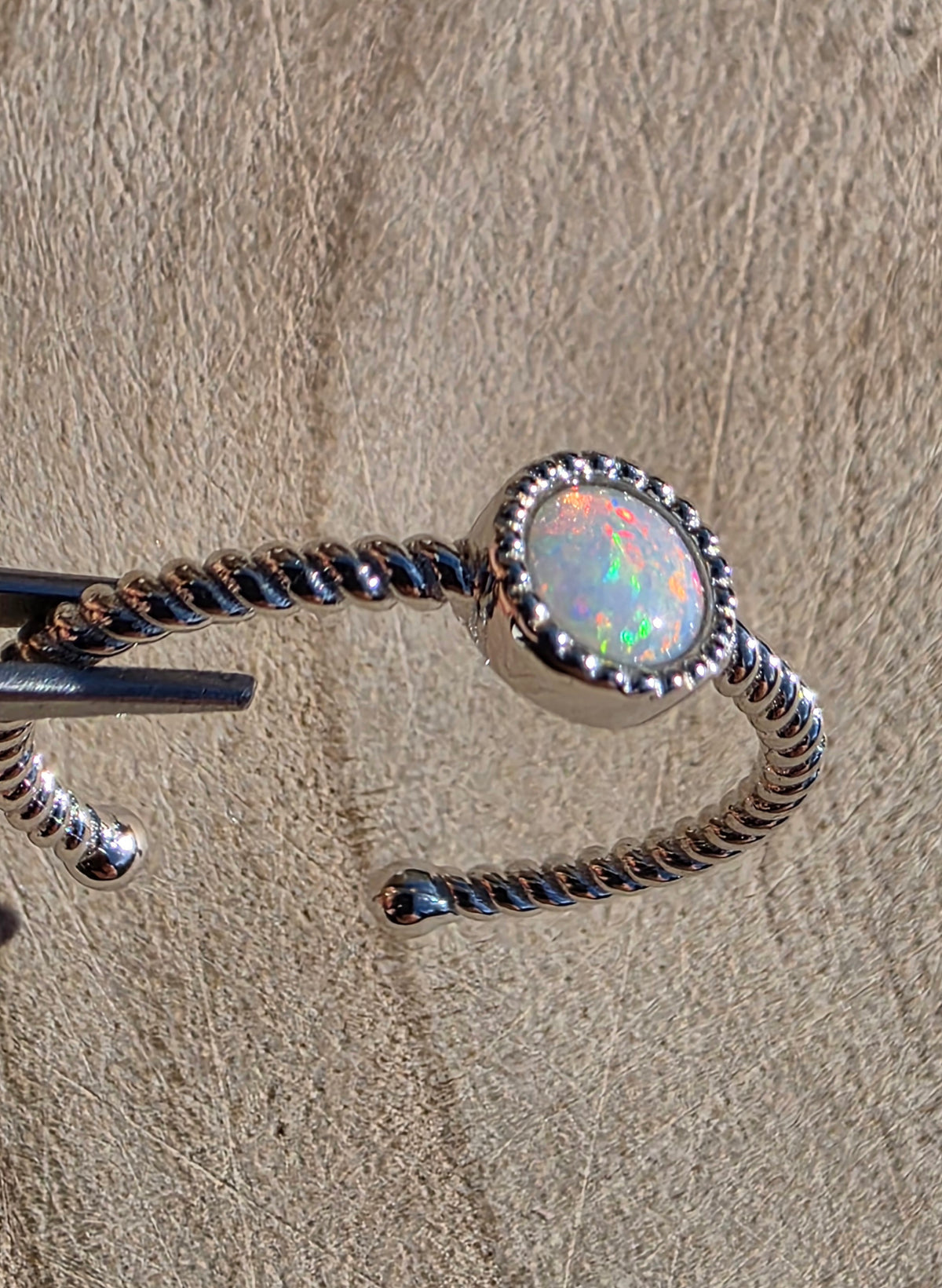 A silver-toned twisted open cuff ring featuring an oval white opal with iridescent multicolour flashes, photographed close up against a textured beige surface.