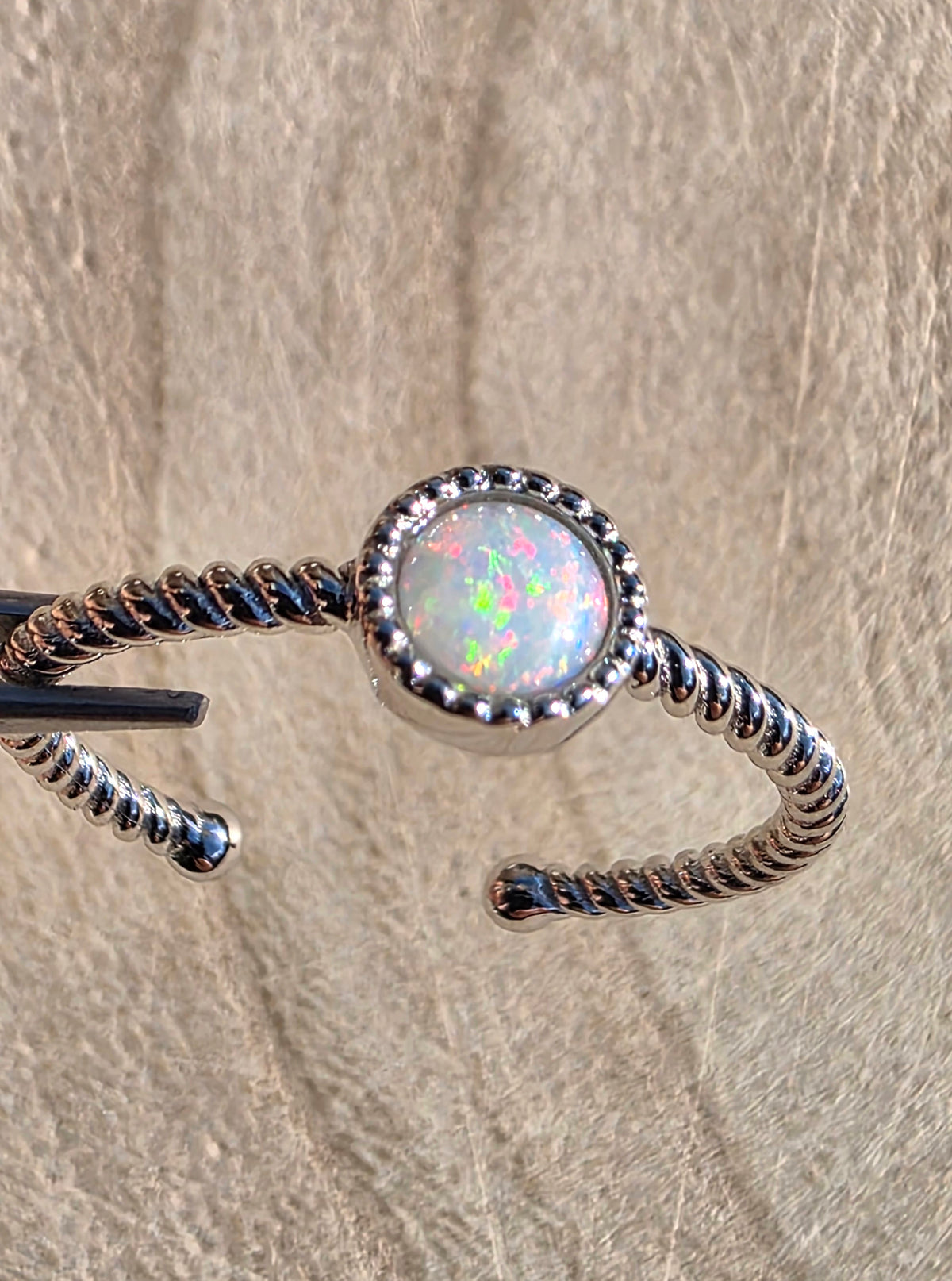 A silver twisted open cuff ring featuring a round white opal centre stone with iridescent flashes, displayed against a textured beige background.