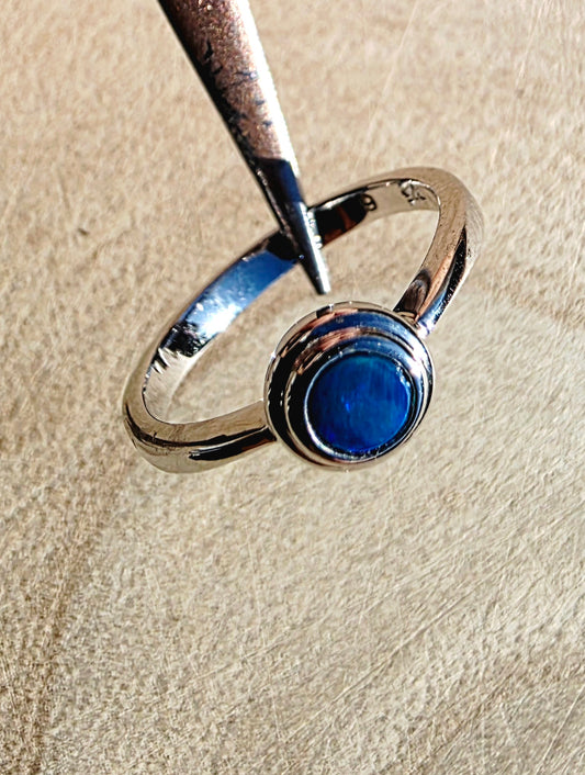 A silver-toned ring with a round deep blue cabochon opal, held by metal tweezers against a textured beige background.