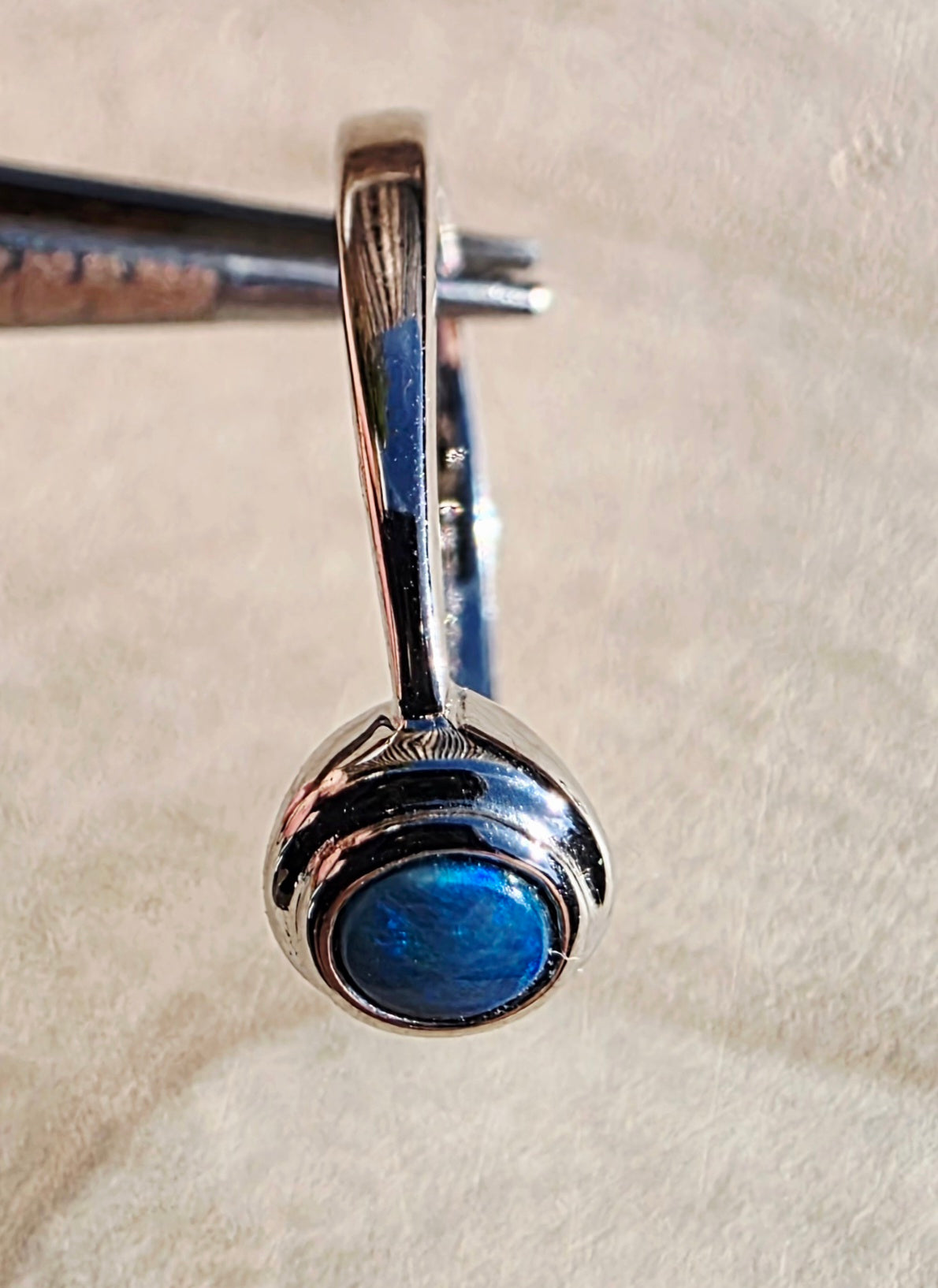 A sterling silver ring with a round blue iridescent opal gemstone, held in forceps against a textured beige background.