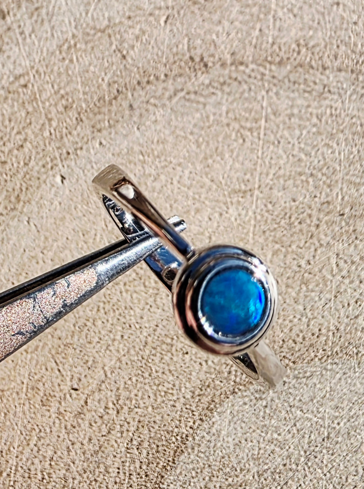 A silver ring with a round blue iridescent opal held by metal tweezers against a textured beige background.
