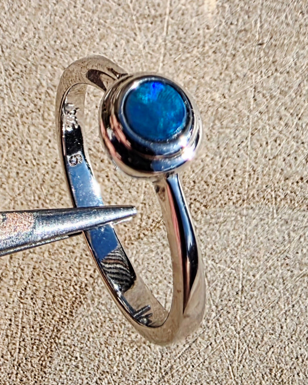 A sterling silver ring with a round blue opal gemstone, held by forceps against a textured beige background.