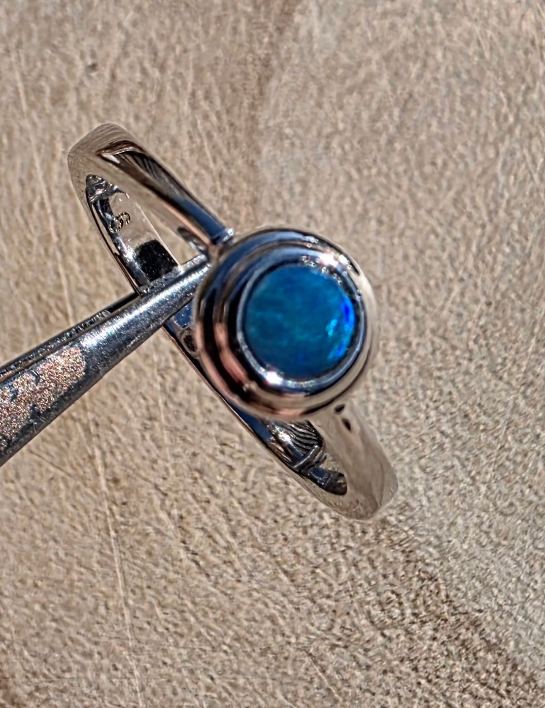 A sterling silver ring with a round, bezel-set blue opal gemstone, held by forceps against a textured beige leather background.