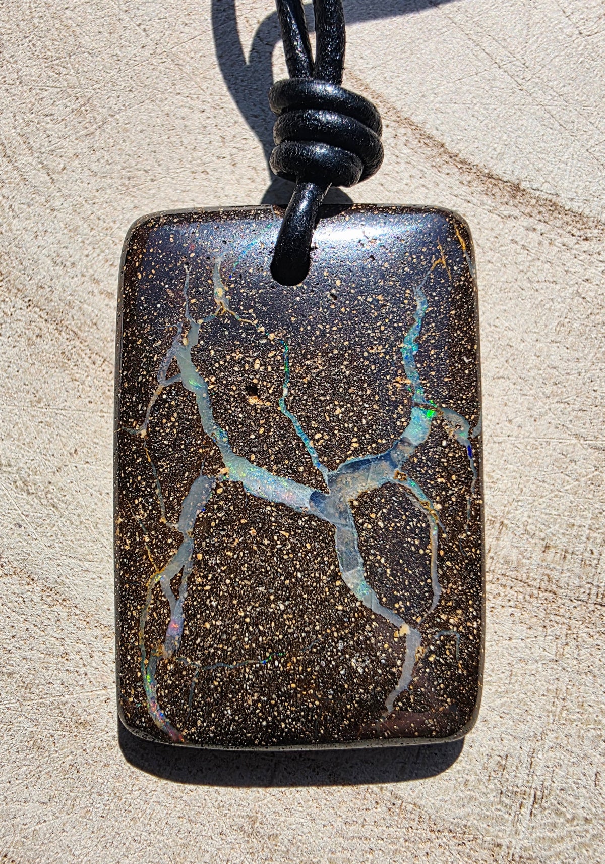 A rectangular dark brown opal pendant with branching iridescent white and blue veins, suspended from a black cord on a sandy beige background.
