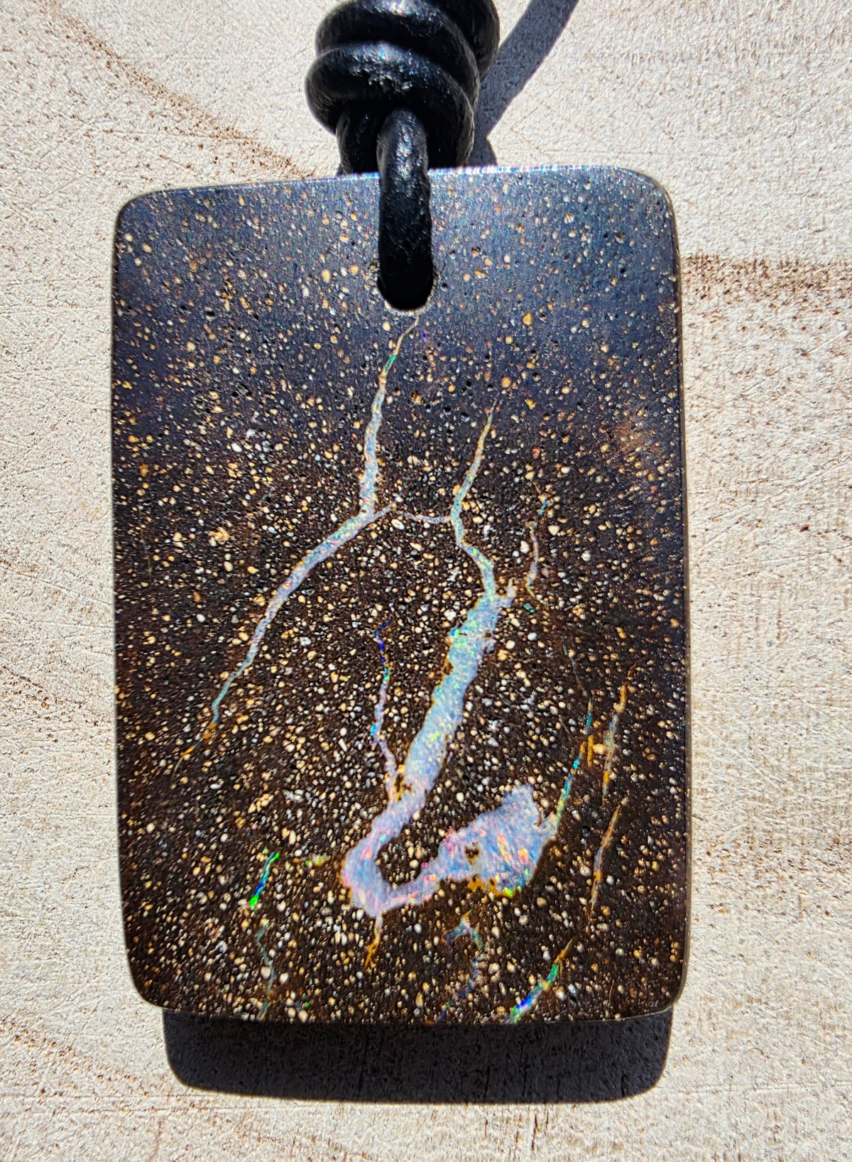 A rectangular black stone pendant with a dark gold speckled background and vibrant white, blue, and iridescent opal veins, hanging from a black cord on a light beige textured surface.