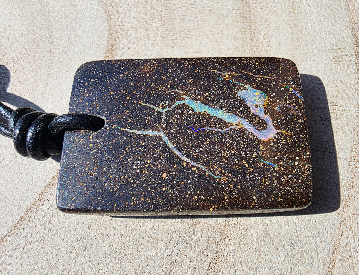 A rectangular black stone pendant with gold flecks and iridescent blue, purple, and pink veining, attached to a black cord, photographed on a light beige textured surface.