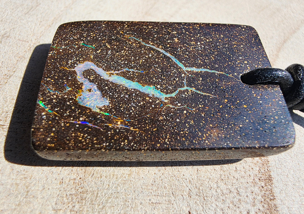 A rectangular black opal pendant with a hole on the right side, featuring a vibrant iridescent opal pattern in white, blue, and gold flecks. The pendant is shown resting on a light beige textured surface with a black cord threaded through the hole.
