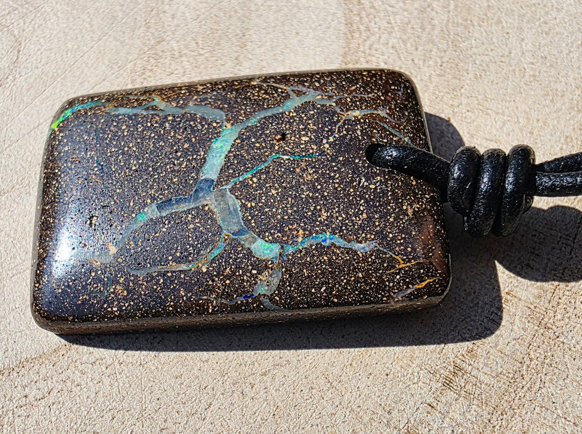 A rectangular dark brown opal pendant with gold flecks and vibrant blue-green veining, attached to a black cord, photographed on a sandy beige surface.