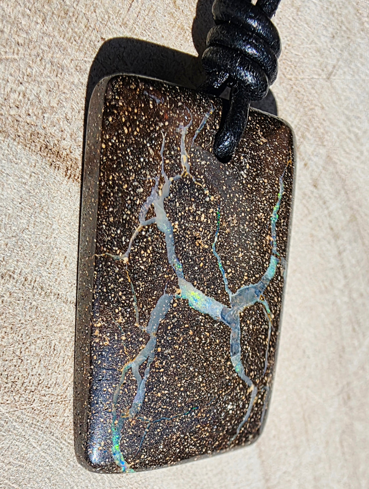 A rectangular dark brown opal pendant with gold flecks and branching light blue-green veining, suspended from a black cord against a light beige textured background.