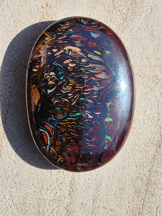 A polished oval cabochon gemstone with a highly iridescent, multicoloured pattern of orange, blue, green, and purple over a dark base, resting on a textured beige surface.