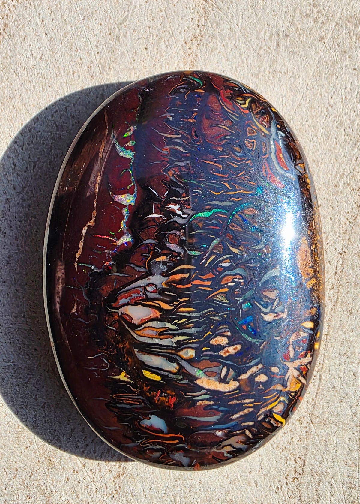 A polished oval opal gemstone with swirling iridescent patterns in red, brown, blue, green, and gold, photographed on a light beige textured surface.