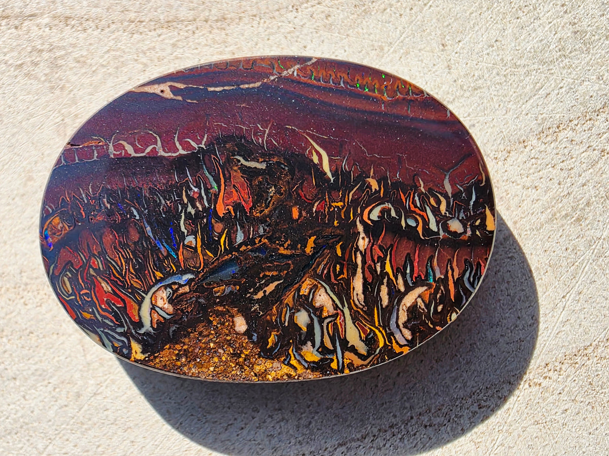 An oval resin cabochon featuring a highly detailed, fiery abstract design with orange, yellow, red, and black swirls, resting on a textured beige stone surface.